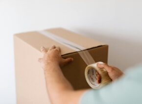 Hands sealing a cardboard box with tape, prepping for a move or delivery indoors.