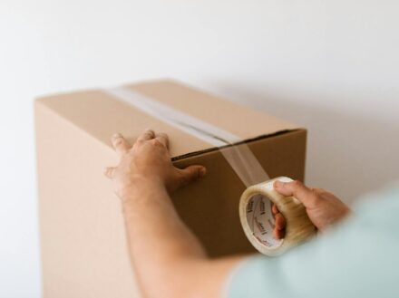 Hands sealing a cardboard box with tape, prepping for a move or delivery indoors.